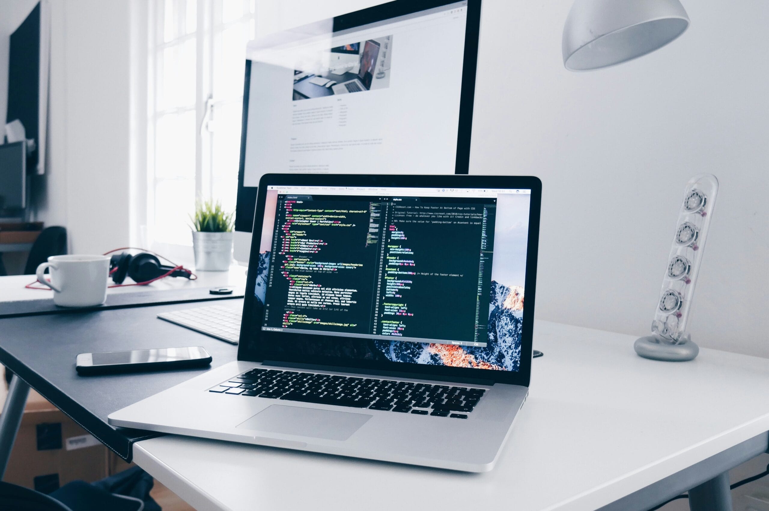 A laptop displaying code on a modern desk with a monitor and office accessories.