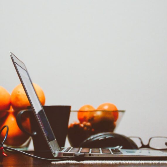 A laptop beside a red mug and a bowl of oranges on a wooden table.