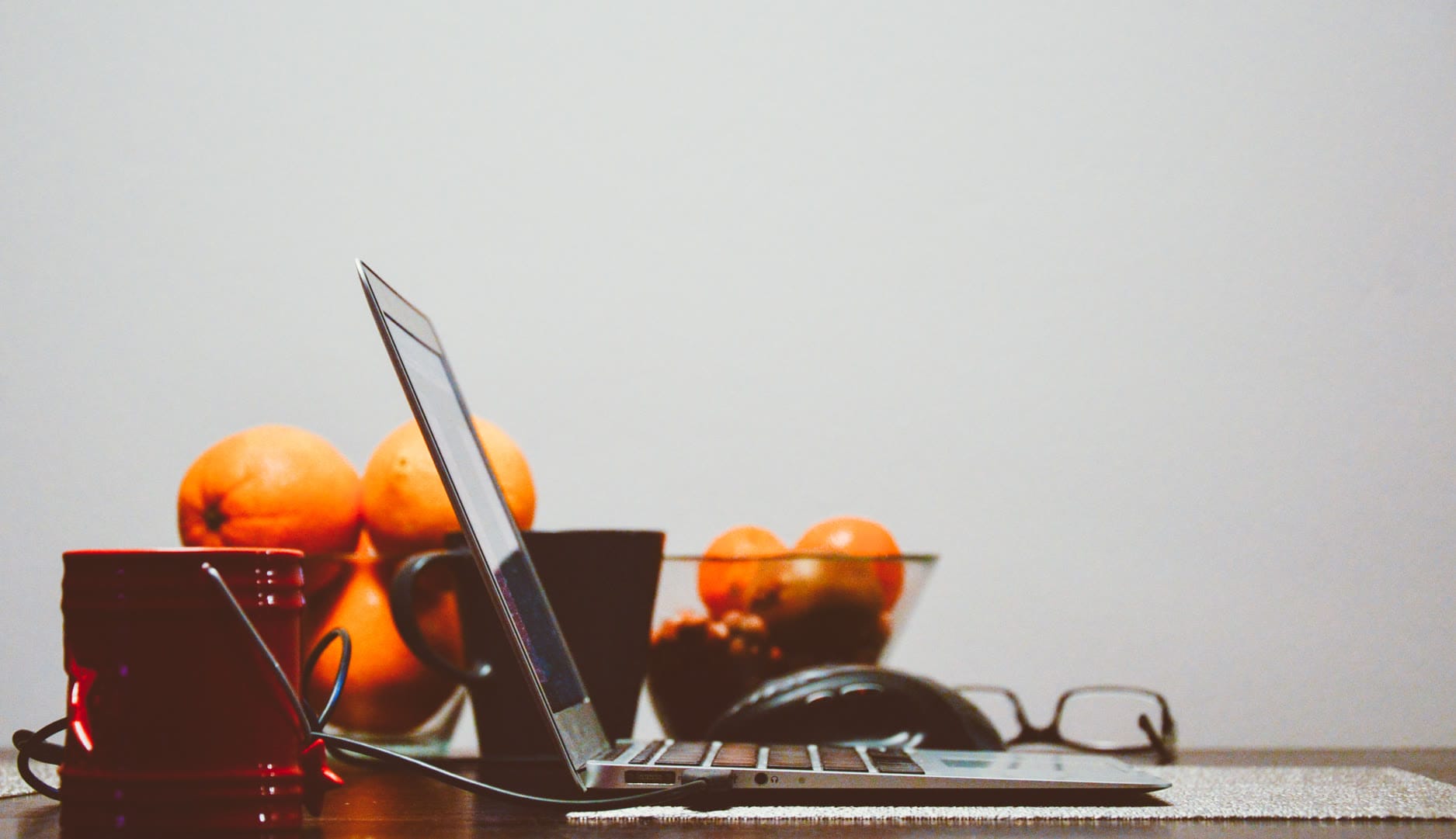 A laptop beside a red mug and a bowl of oranges on a wooden table.