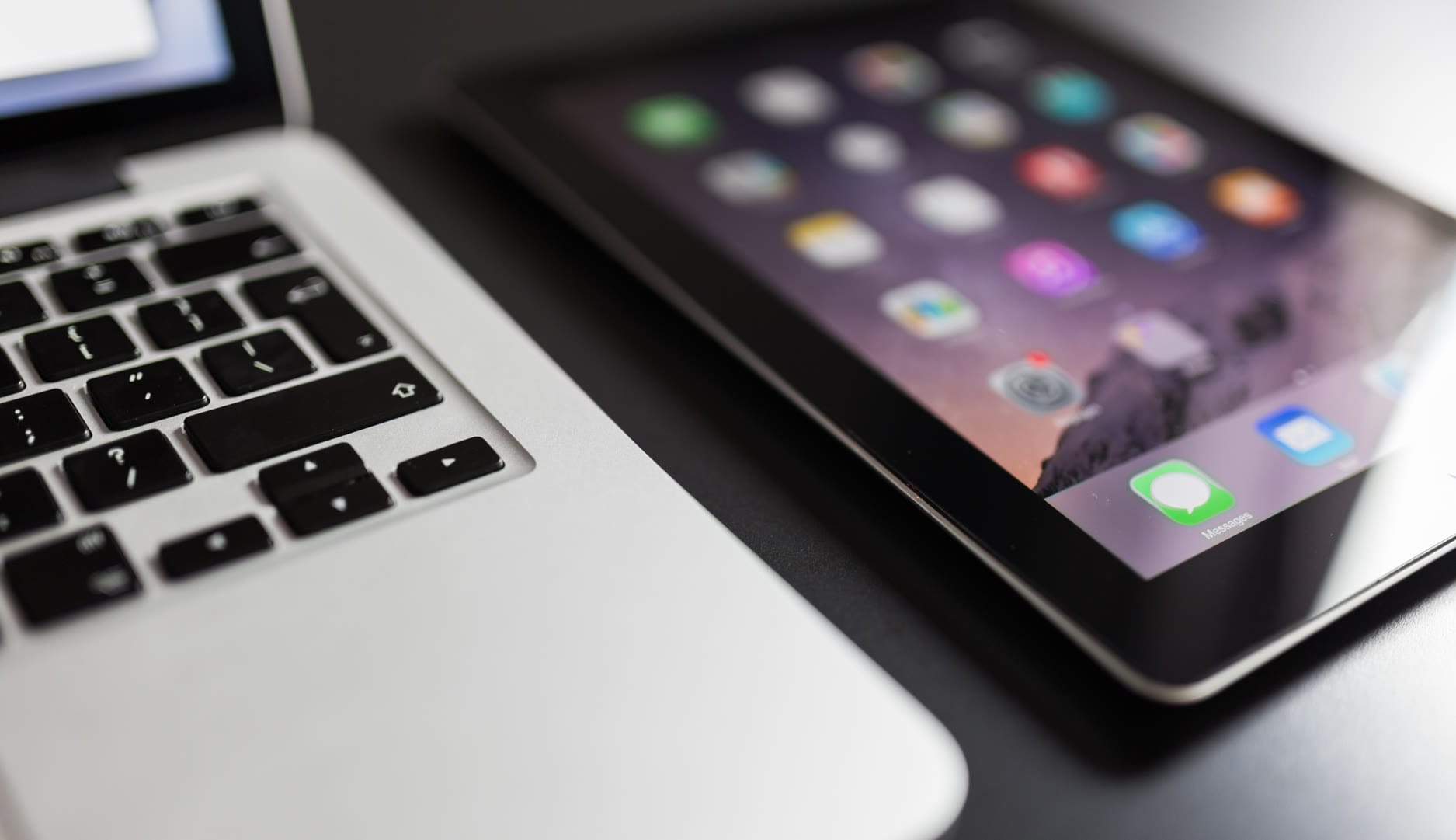 A laptop keyboard next to a tablet displaying app icons on a dark surface.