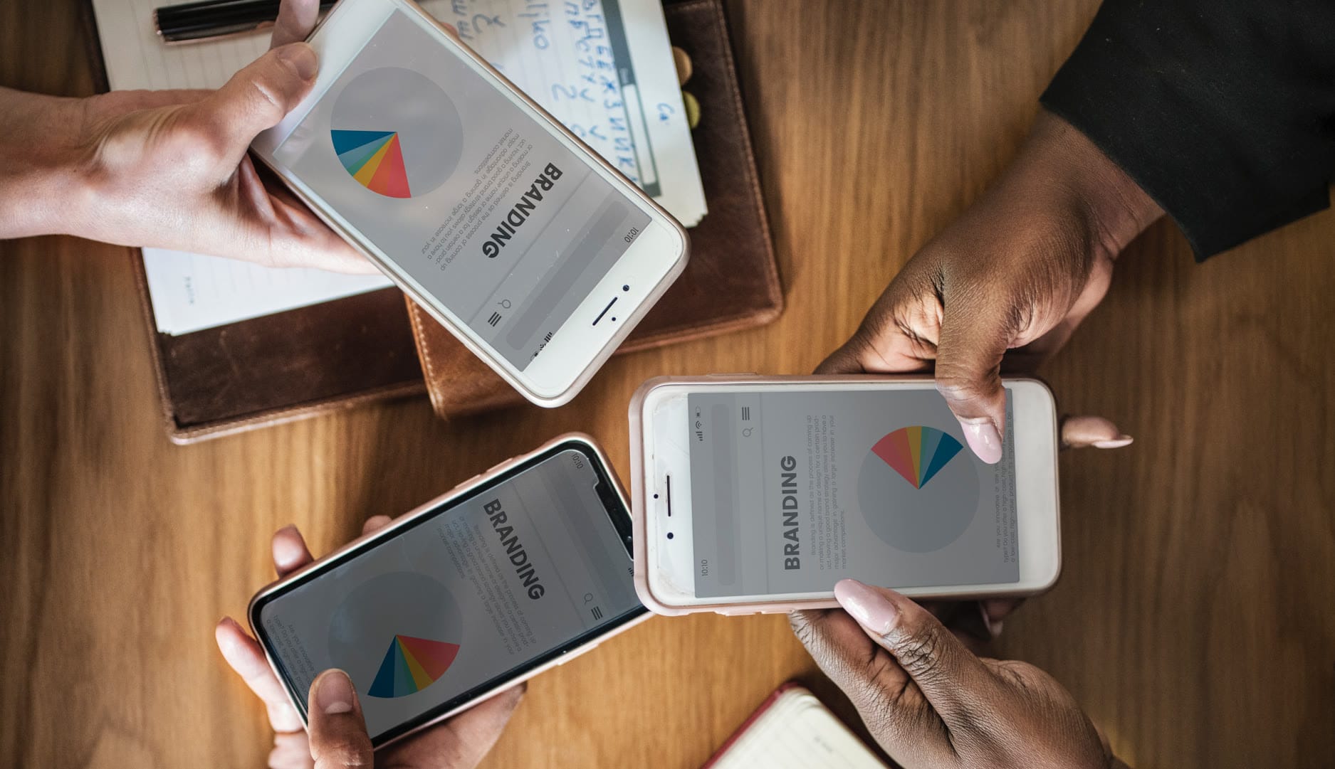 Three hands holding smartphones displaying branding pie charts on a wooden table.